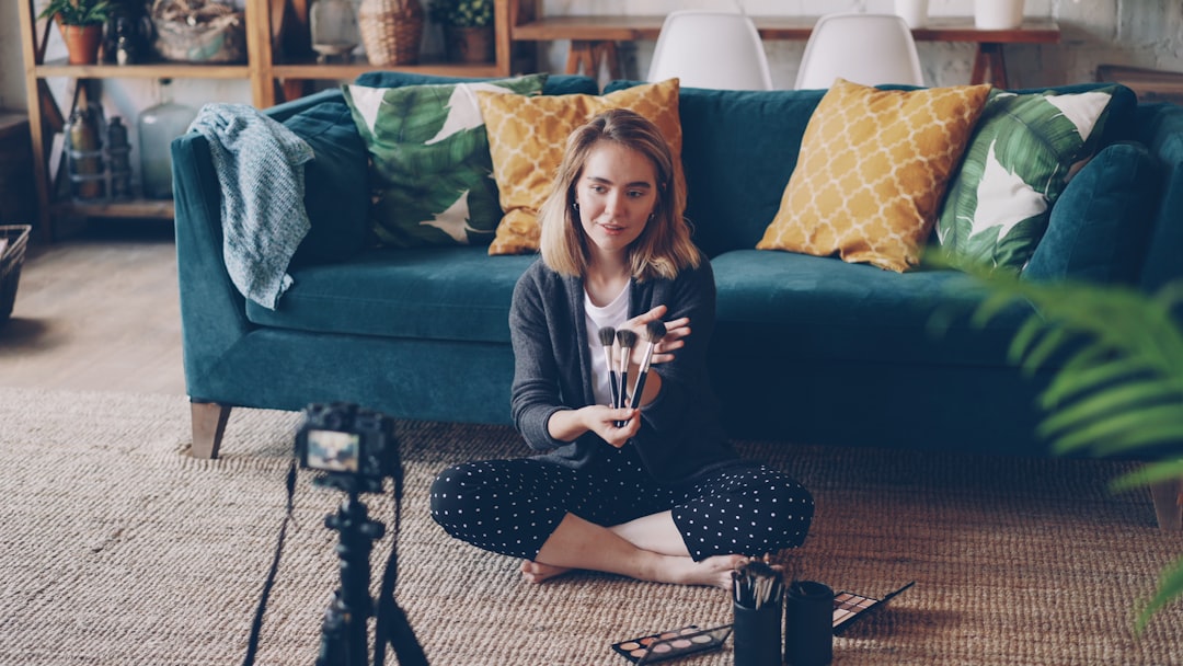 Cheerful girl vlogger is recording video about make-up, cosmetics and tools for her internet videolog using camera. Woman is sitting on floor, showing beauty products and speaking.