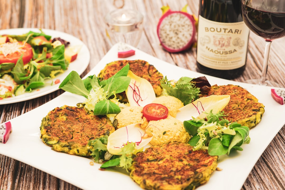 Crispy zucchini fritters with hummus, fresh mixed greens and chicory, served with red wine and dragon fruit. A creative Mediterranean vegetarian restaurant dish — perfect for restaurant menus, food blogs and gastronomy websites.